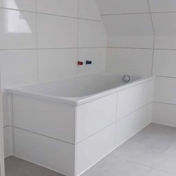 Sławomir Kamiński - Biała, zabudowana wanna w łazience na poddaszu, wykończona płytkami w odcieniach bieli i szarości. Widoczne baterie z oznaczeniem ciepłej i zimnej wody.