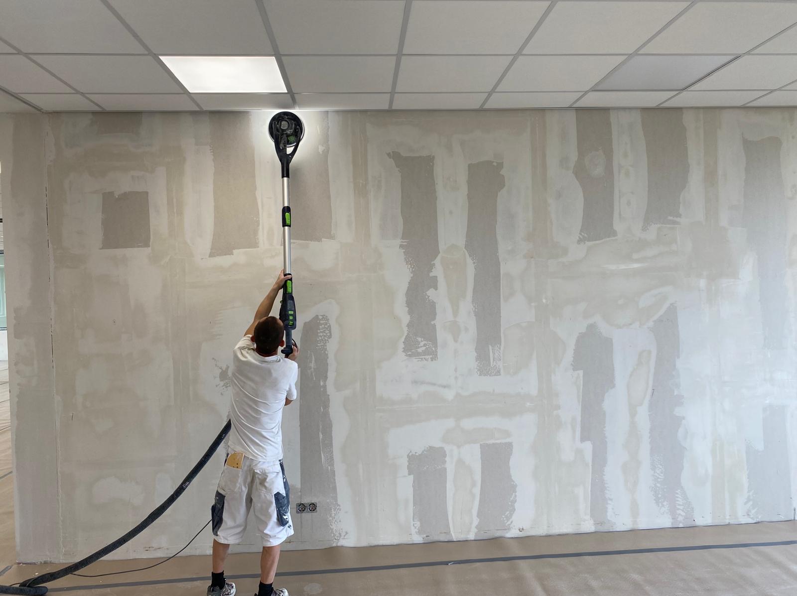 Pracownik szlifuje ścianę za pomocą szlifierki z odkurzaczem w pomieszczeniu biurowym. Widoczne ślady po szpachlowaniu i przygotowaniu pod malowanie. Sufit kasetonowy.