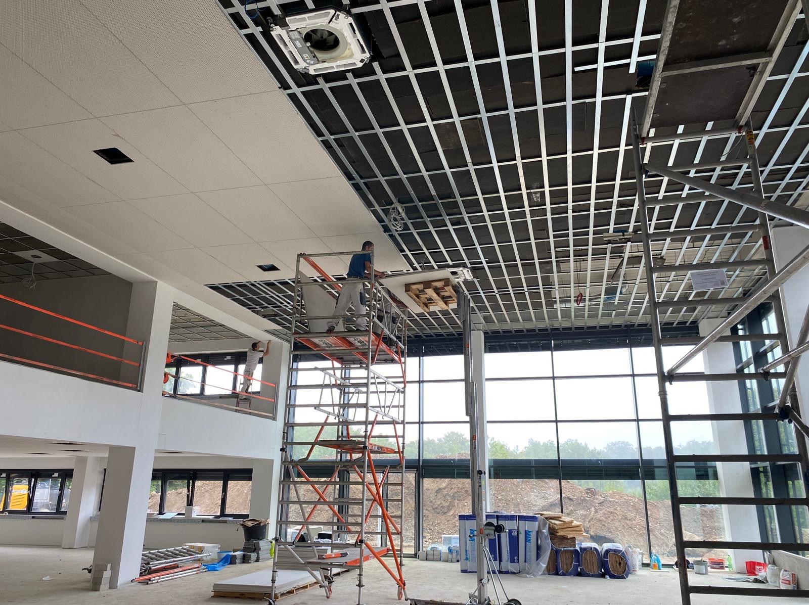 Montaż sufitu podwieszanego w biurze: pracownik na rusztowaniu instaluje panele sufitowe w metalowej konstrukcji. Widoczne okna i materiały budowlane.