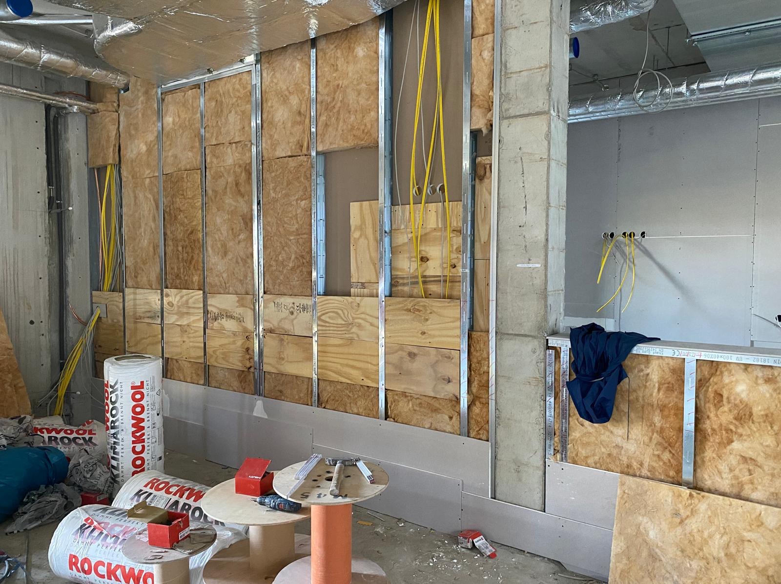 Montaż izolacji z wełny mineralnej i płyt gipsowo-kartonowych na ścianie. Widoczne profile, instalacja elektryczna, narzędzia i materiały budowlane na podłodze.