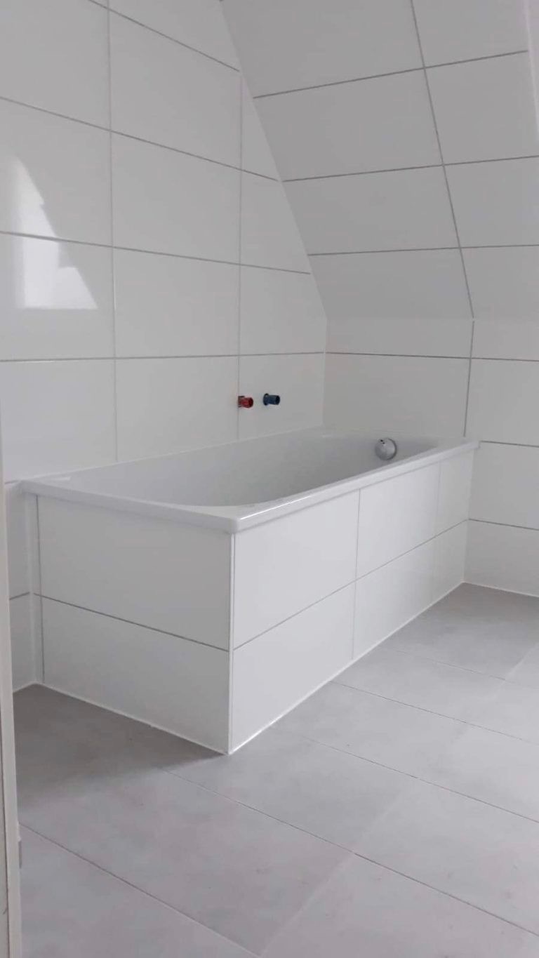 Biała, zabudowana wanna w łazience na poddaszu, wykończona płytkami w odcieniach bieli i szarości. Widoczne baterie z oznaczeniem ciepłej i zimnej wody.