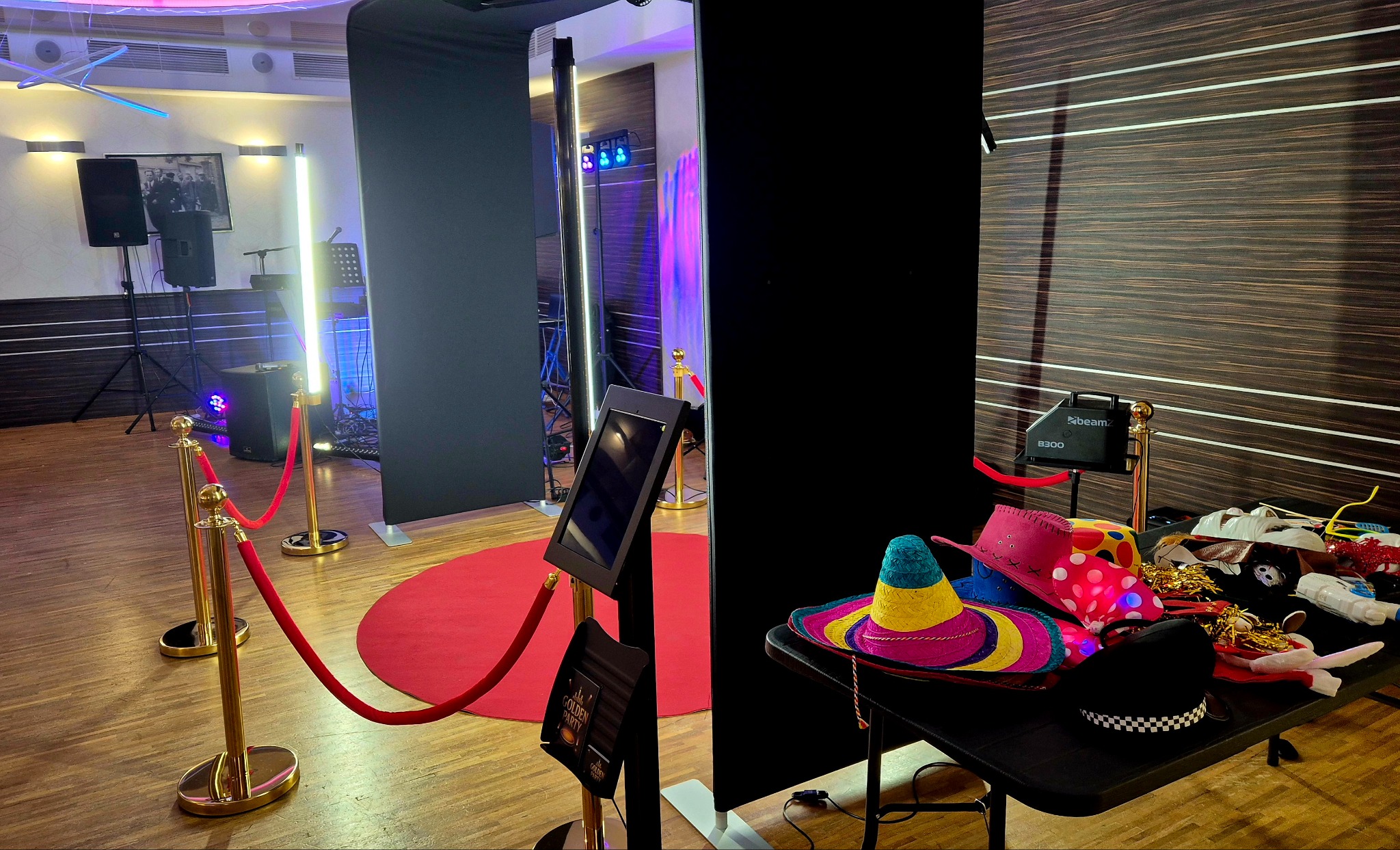Kompletna fotobudka z rekwizytami: kolorowe kapelusze, maski, czerwony dywan i złote słupki odgradzające. Gotowa do użycia na imprezie.