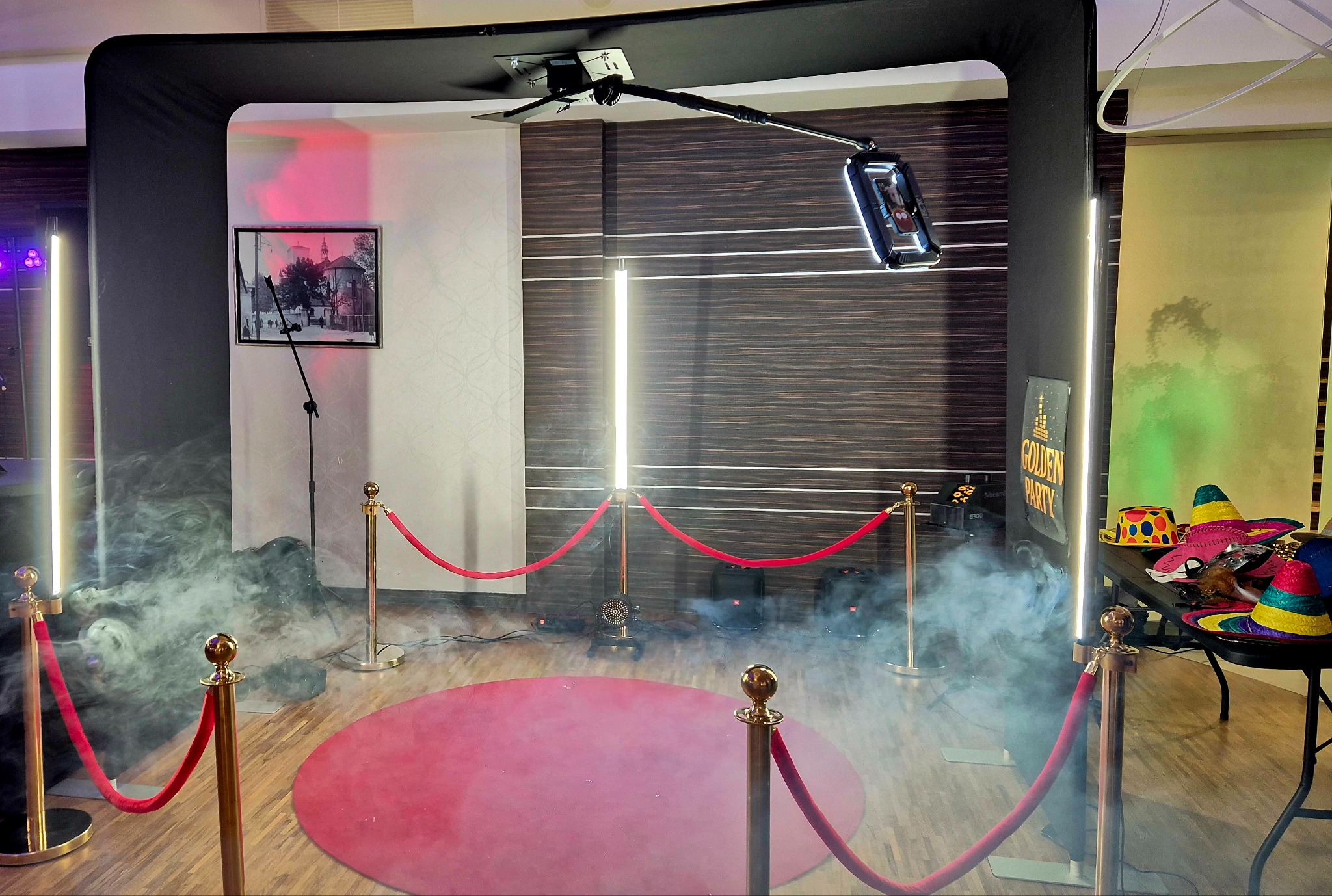 Nowoczesna fotobudka z czerwonym dywanem, oświetleniem LED i rekwizytami (kapelusze). Czarny stelaż, automat do zdjęć na wysięgniku. Dekoracja z efektem dymu.