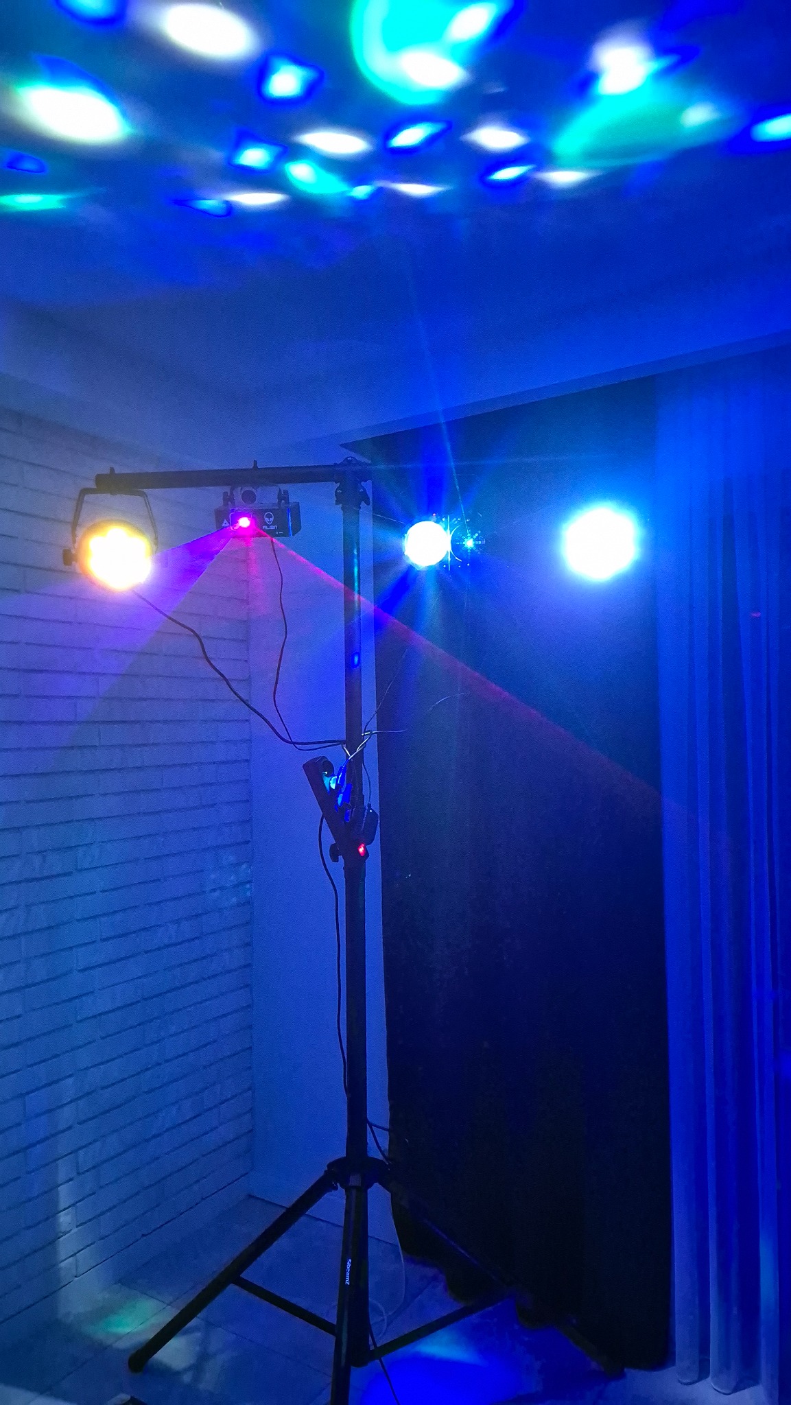 Stojak oświetleniowy z reflektorami i laserem, rzucający kolorowe światła na sufit i ścianę z cegły. Efekt dyskotekowy, niebieska poświata.