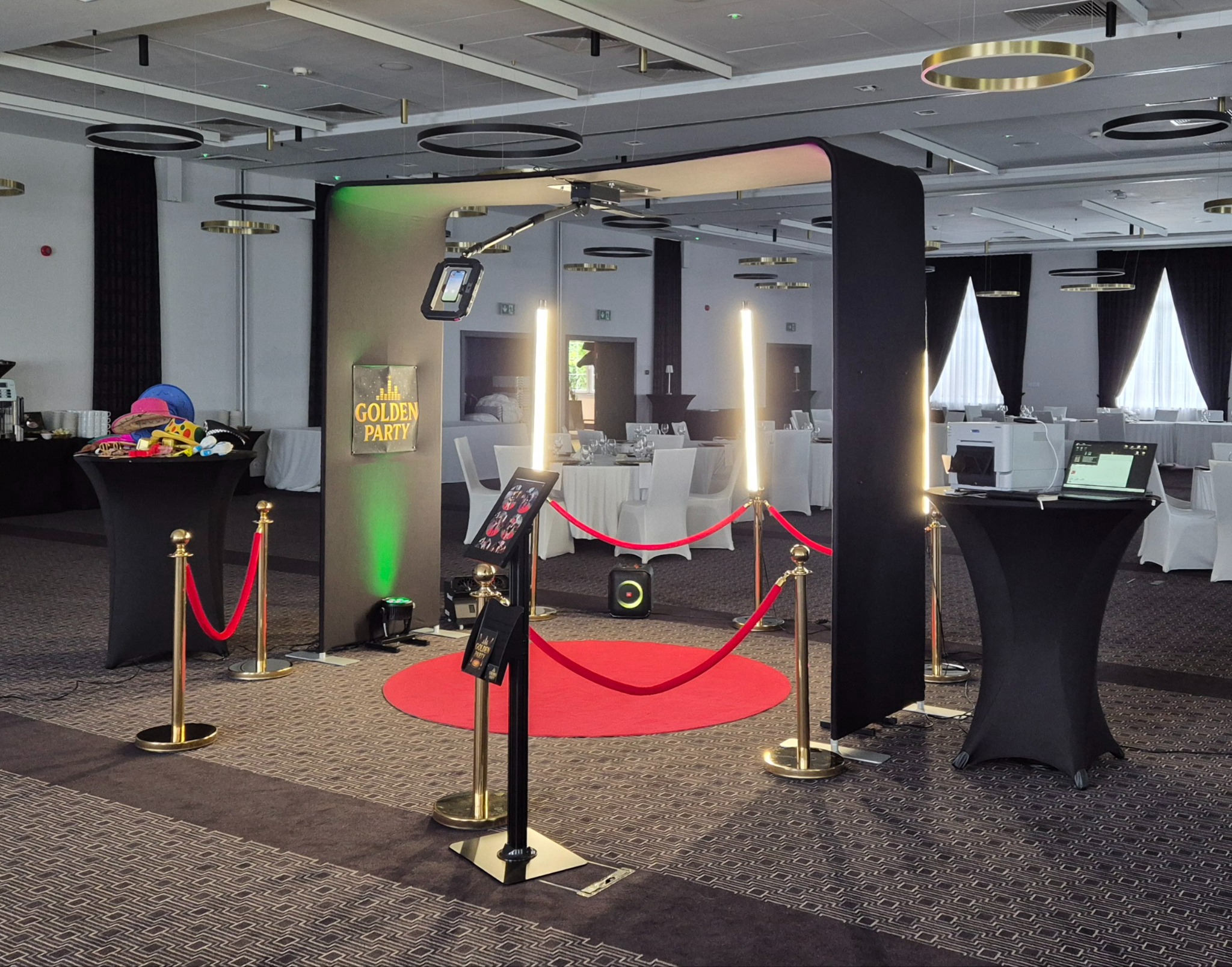 Elegancka fotobudka 'Golden Party' z czerwonym dywanem i złotymi słupkami w sali bankietowej. Oświetlenie LED i rekwizyty na stole dodają uroku.