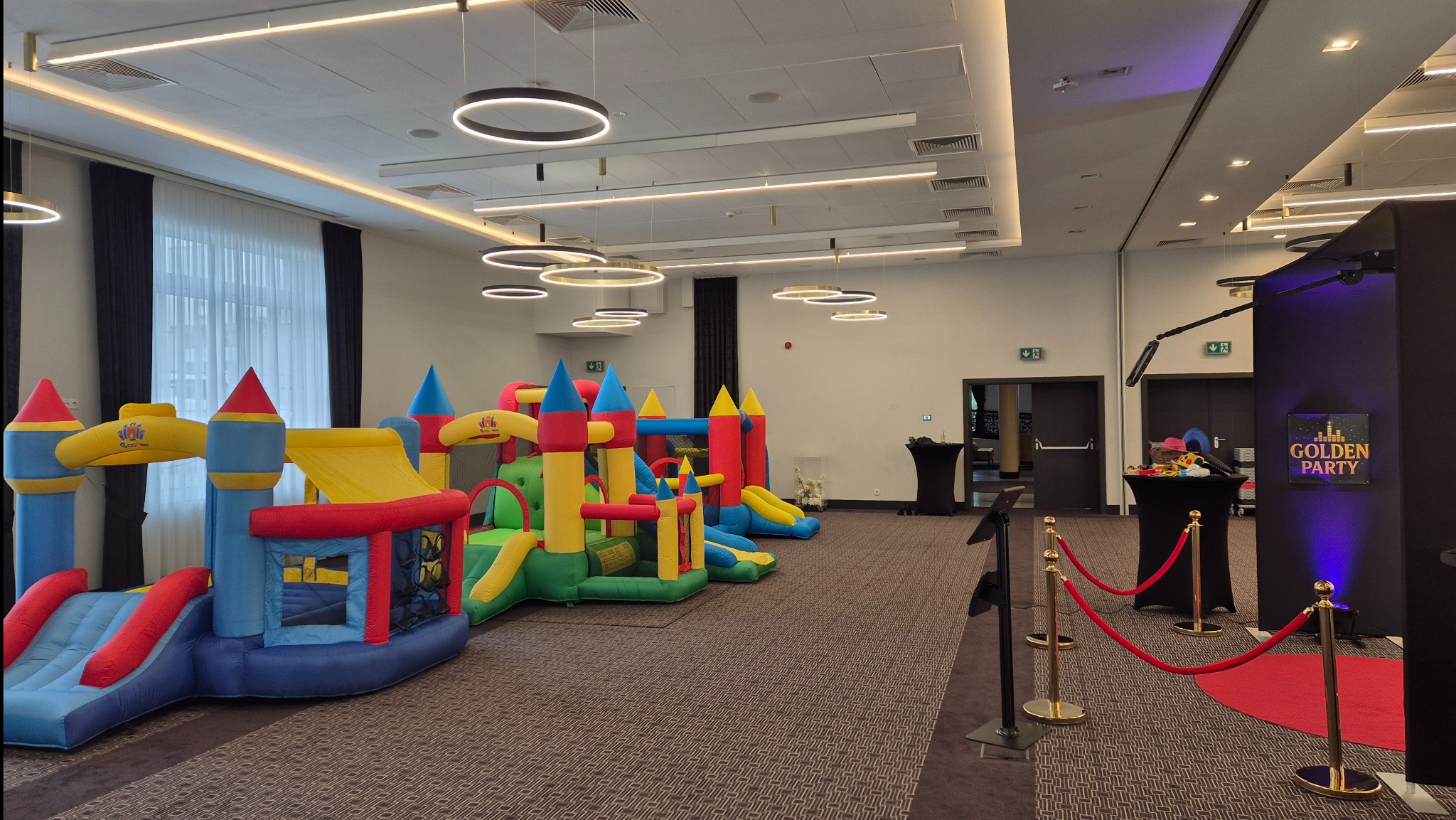 Kolorowe dmuchańce zamki w przestronnej sali z fotobudką Golden Party. Ustawione w rzędzie, gotowe do zabawy. Oświetlenie sufitowe, ciemne zasłony i wykładzina.