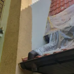 Fragment elewacji domu w trakcie malowania: część ściany pomalowana na biało, reszta w odcieniach beżu. Widoczne zabezpieczenia folią malarską na dachu.