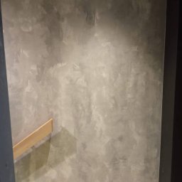 Patryk Siwicki - Ściana po malowaniu dekoracyjnym w odcieniach szarości z widoczną fakturą. Na podłodze narzędzia malarskie i folia ochronna. Widoczny fragment drewnianej listwy.