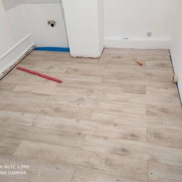 Patryk Siwicki - Jasne panele podłogowe ułożone w pomieszczeniu z białymi ścianami. Widoczna poziomica, śrubokręt i taśma malarska. Etap prac wykończeniowych wnętrza.