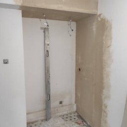 Patryk Siwicki - Przygotowanie ściany do malowania: ściana z nałożoną gładzią, poziomica oparta o ścianę, widoczne kable, podłoga zabezpieczona folią malarską, wiadro z farbą.