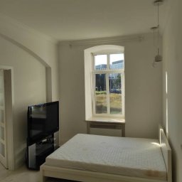 Patryk Siwicki - Jasne wnętrze pokoju po malowaniu: białe ściany, łóżko z materacem, okno z widokiem na zieleń, telewizor i lampy wiszące. Minimalistyczny styl.