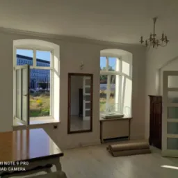 Jasne wnętrze z drewnianą podłogą i dużymi oknami z widokiem na budynek biurowy. Świeżo pomalowane ściany, w rogu pokoju stoi antyczna komoda.