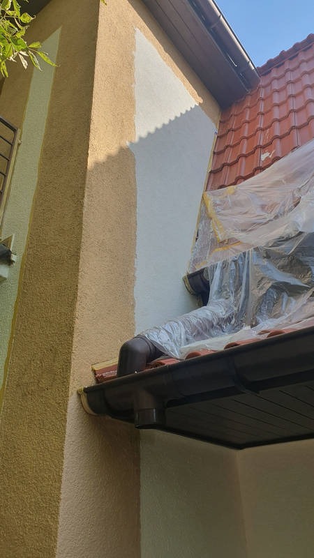 Fragment elewacji domu w trakcie malowania: część ściany pomalowana na biało, reszta w odcieniach beżu. Widoczne zabezpieczenia folią malarską na dachu.