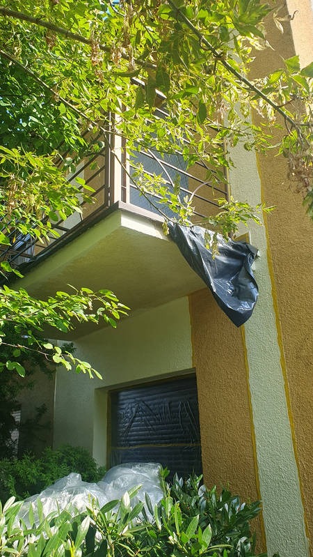 Przygotowania do prac na elewacji: okno i balkon zabezpieczone folią malarską, widoczne świeże, jasne fragmenty tynku. Częściowo przysłonięte przez zieleń.