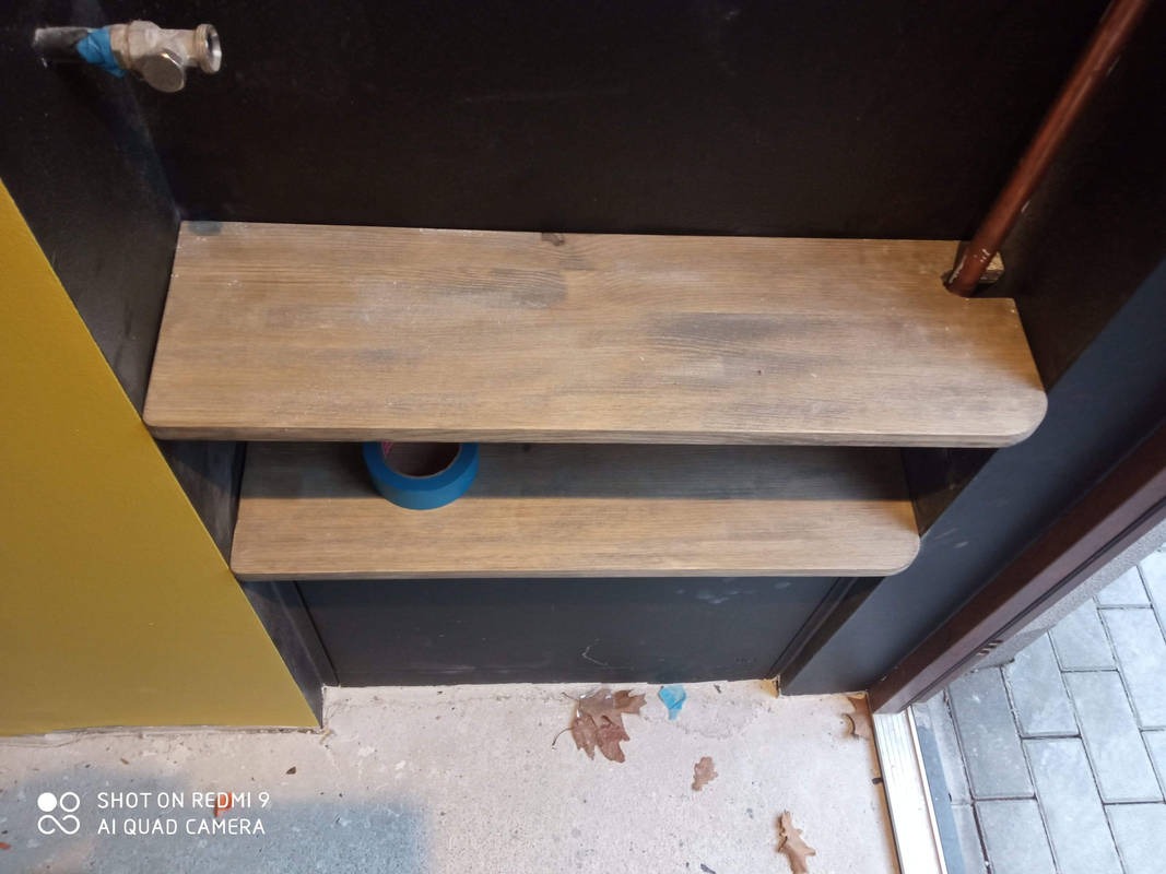 Dwie drewniane półki wnękowe, pomalowane na brązowo, z zaokrąglonymi krawędziami. Na dolnej półce leży niebieska taśma malarska. Żółta ściana z lewej.