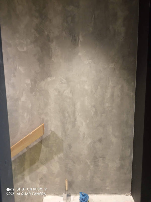 Ściana po malowaniu dekoracyjnym w odcieniach szarości z widoczną fakturą. Na podłodze narzędzia malarskie i folia ochronna. Widoczny fragment drewnianej listwy.