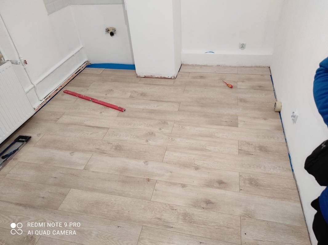 Jasne panele podłogowe ułożone w pomieszczeniu z białymi ścianami. Widoczna poziomica, śrubokręt i taśma malarska. Etap prac wykończeniowych wnętrza.