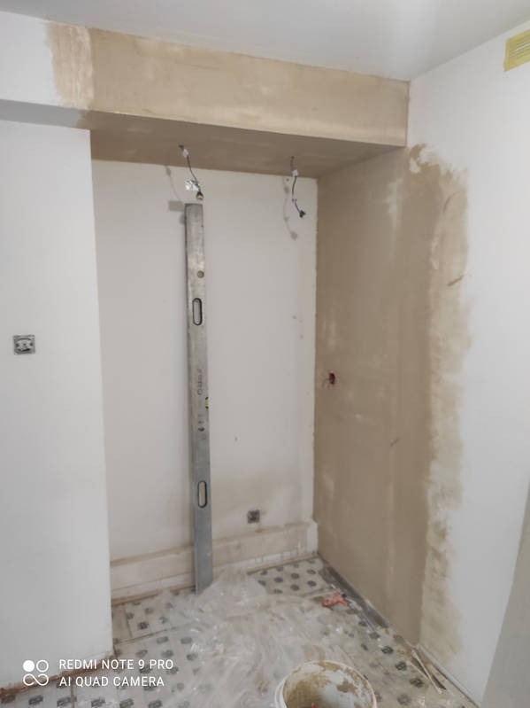 Przygotowanie ściany do malowania: ściana z nałożoną gładzią, poziomica oparta o ścianę, widoczne kable, podłoga zabezpieczona folią malarską, wiadro z farbą.