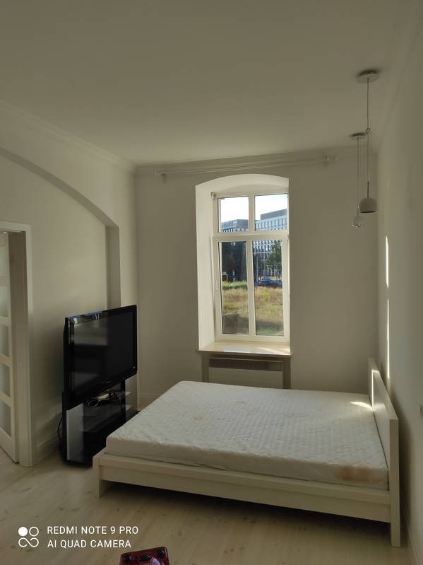 Jasne wnętrze pokoju po malowaniu: białe ściany, łóżko z materacem, okno z widokiem na zieleń, telewizor i lampy wiszące. Minimalistyczny styl.