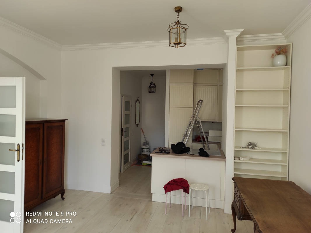 Jasne wnętrze po malowaniu: białe ściany, sztukateria, drewniane meble, drabina malarska. Widoczny efekt odświeżonego pomieszczenia.