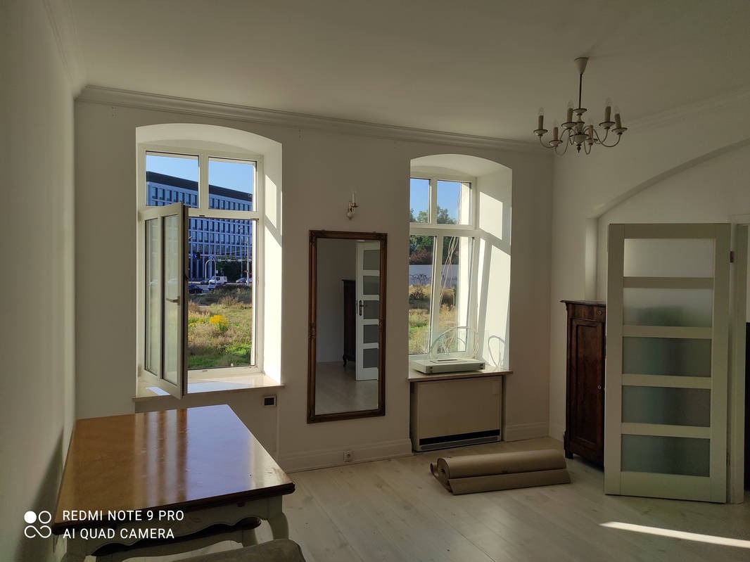 Jasne wnętrze z drewnianą podłogą i dużymi oknami z widokiem na budynek biurowy. Świeżo pomalowane ściany, w rogu pokoju stoi antyczna komoda.