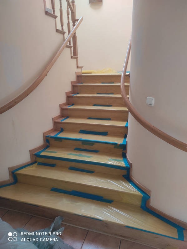 Schody drewniane zabezpieczone folią i taśmą malarską, przygotowane do malowania. Widoczne beżowe ściany i fragment balustrady.