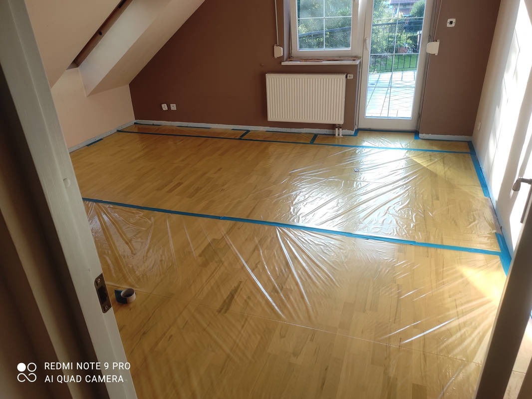 Pokój na poddaszu zabezpieczony folią i taśmą malarską przed malowaniem ścian. Widoczny kaloryfer, okno balkonowe i skos dachu. Przygotowania do prac malarskich.