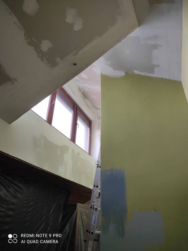 Wnętrze w trakcie prac przygotowawczych: szpachlowanie sufitu i ścian, widoczna drabina i okno. Fragment ściany z próbkami kolorów farb.