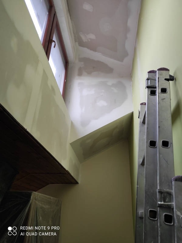 Wnętrze w trakcie prac wykończeniowych: ściany z nałożoną gładzią, okno z brązową ramą, drabina oparta o ścianę. Widoczne ślady szpachlowania na suficie.