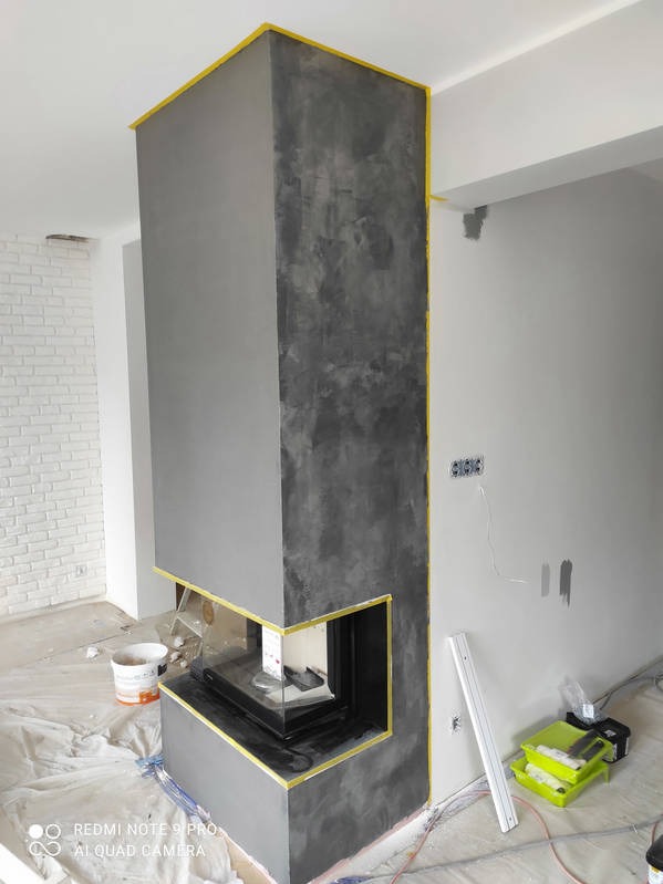 Wykończony kominek w salonie we Wrocławiu, z szarą, dekoracyjną powłoką imitującą beton, zabezpieczony taśmą malarską. Widoczne narzędzia i przygotowane podłoże.