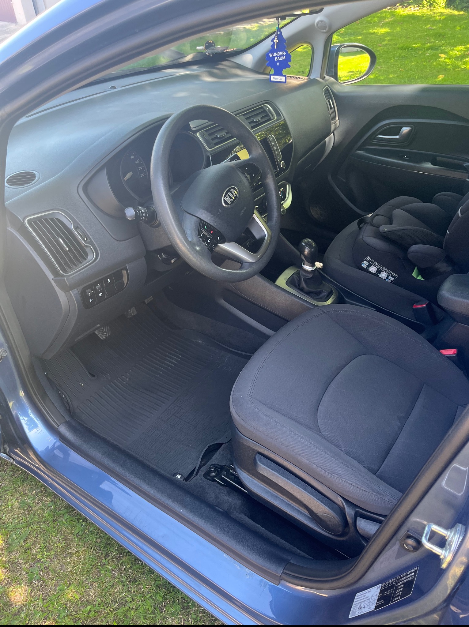 Wnętrze niebieskiego samochodu Kia z czarną tapicerką i fotelikiem dziecięcym na tylnym siedzeniu, widok z otwartych drzwi kierowcy.