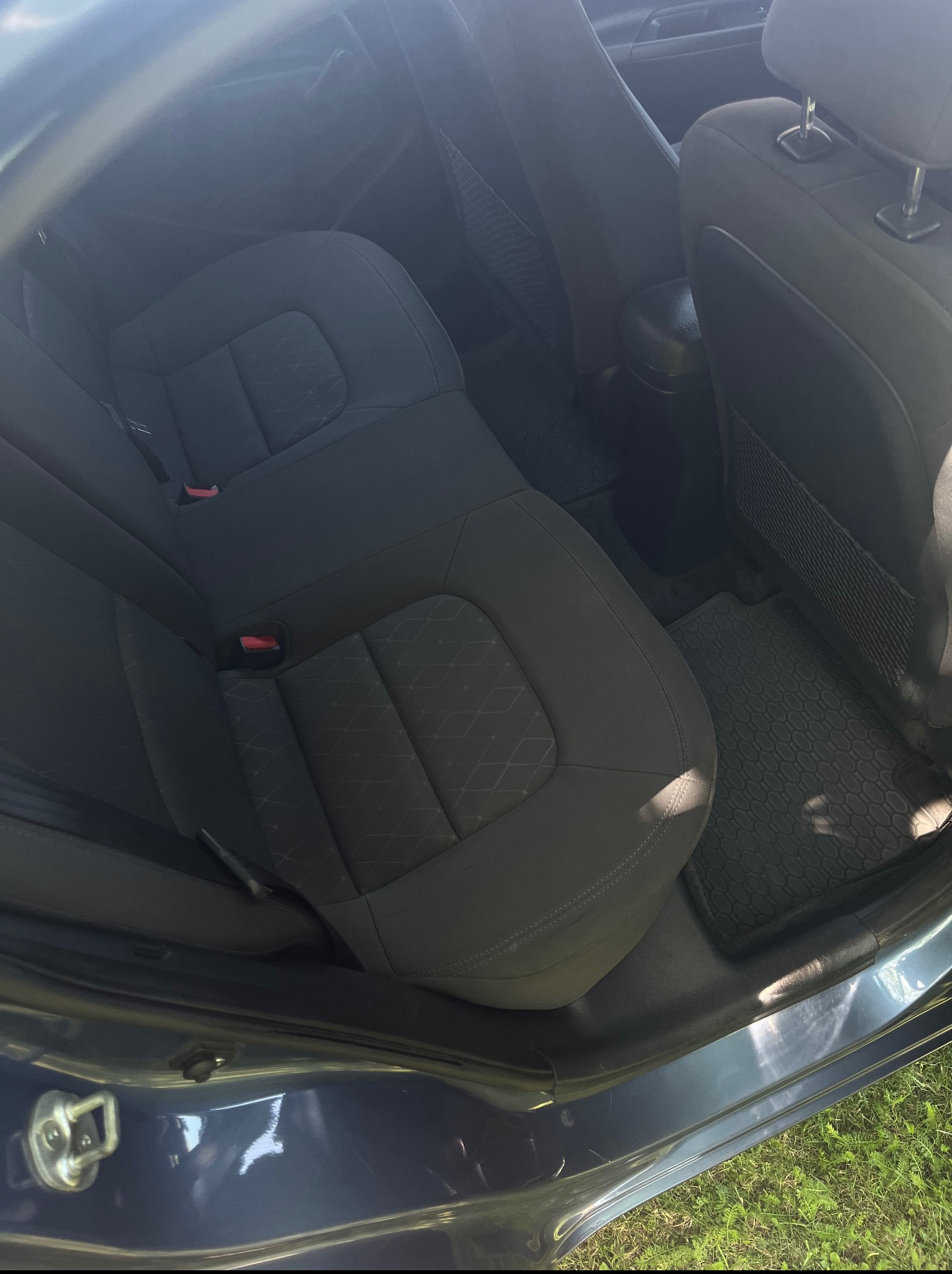 Wnętrze samochodu z ciemną tapicerką. Widoczne tylne siedzenia z zagłówkami i wzorem. Dywaniki gumowe na podłodze. Otwarta rama drzwi auta.