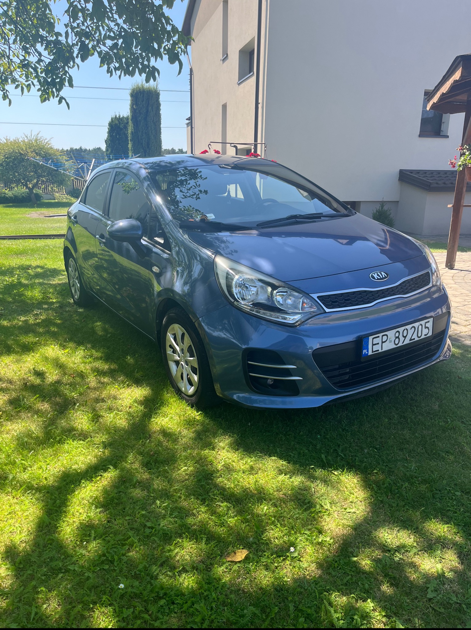 Błękitne auto Kia Rio na trawniku w Wolborzu, z numerem rejestracyjnym EP 89205, w tle dom i drzewa, słoneczny dzień.