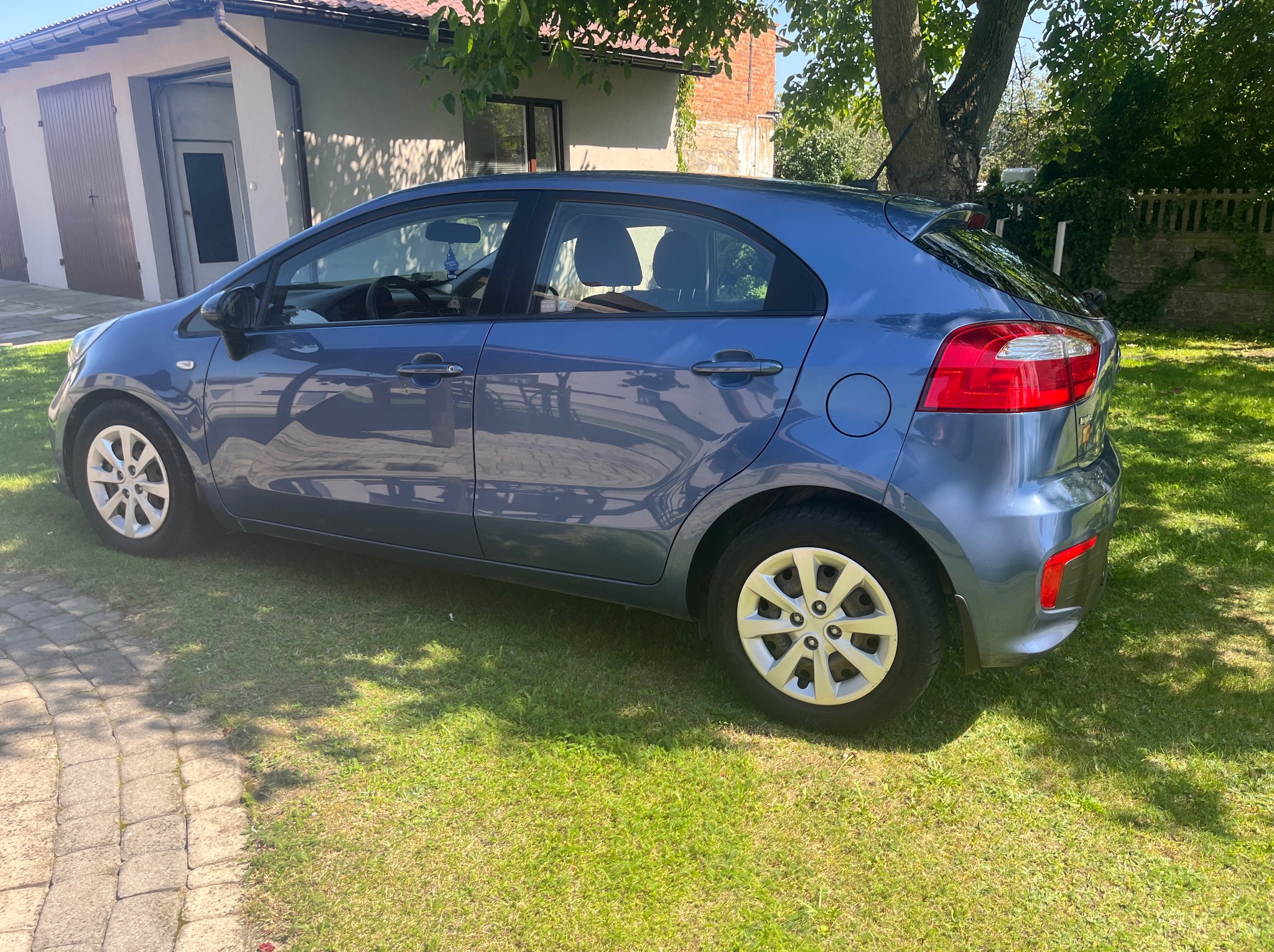 Błękitny samochód Kia Rio zaparkowany na trawniku przed garażem w słoneczny dzień. Widok z boku, cień drzew na trawie. Felgi aluminiowe i tylne światła widoczne.