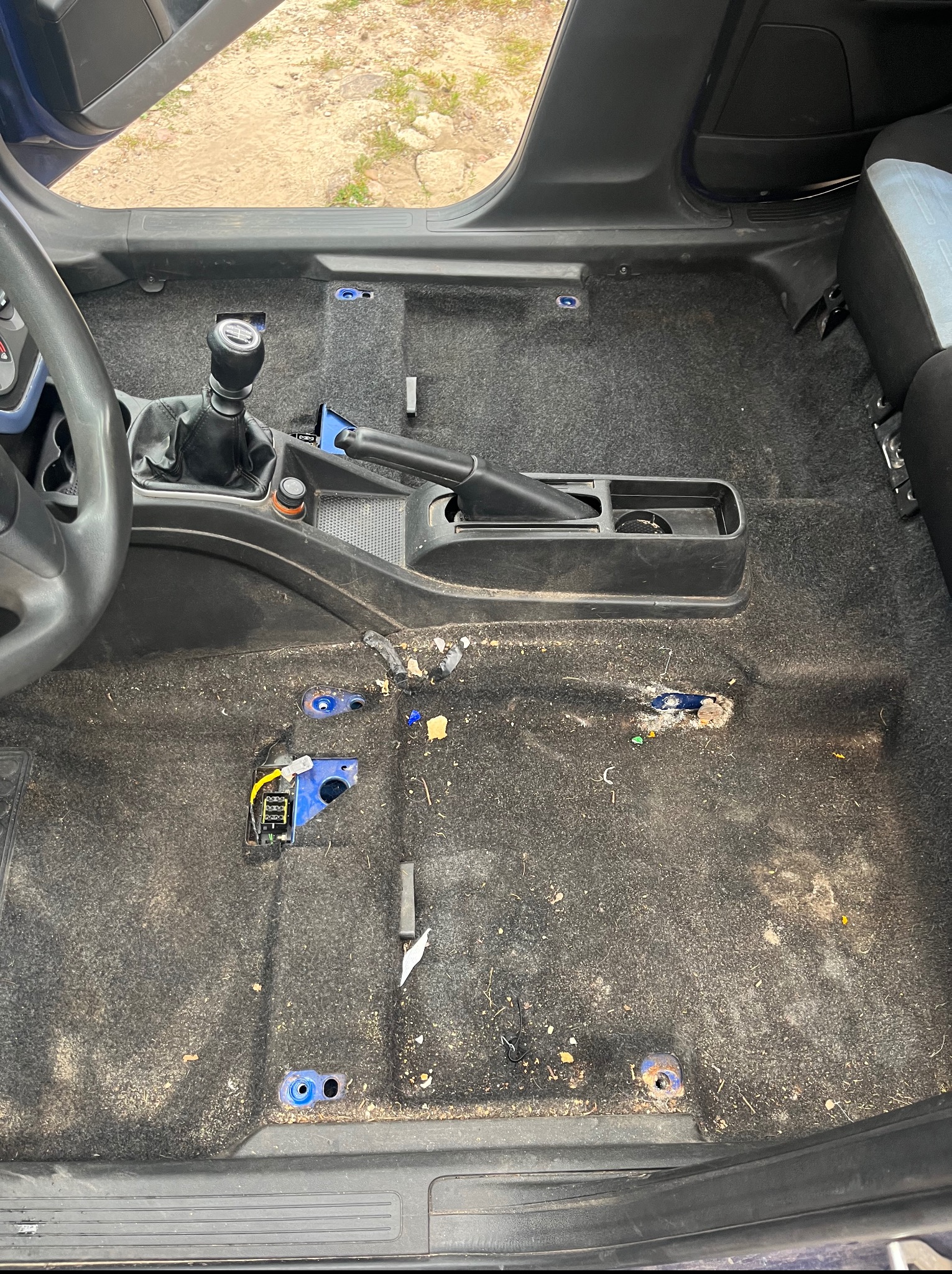 Wnętrze brudnego samochodu przed czyszczeniem: widoczna kierownica, dźwignia zmiany biegów, hamulec ręczny i mocno zabrudzona tapicerka podłogowa.