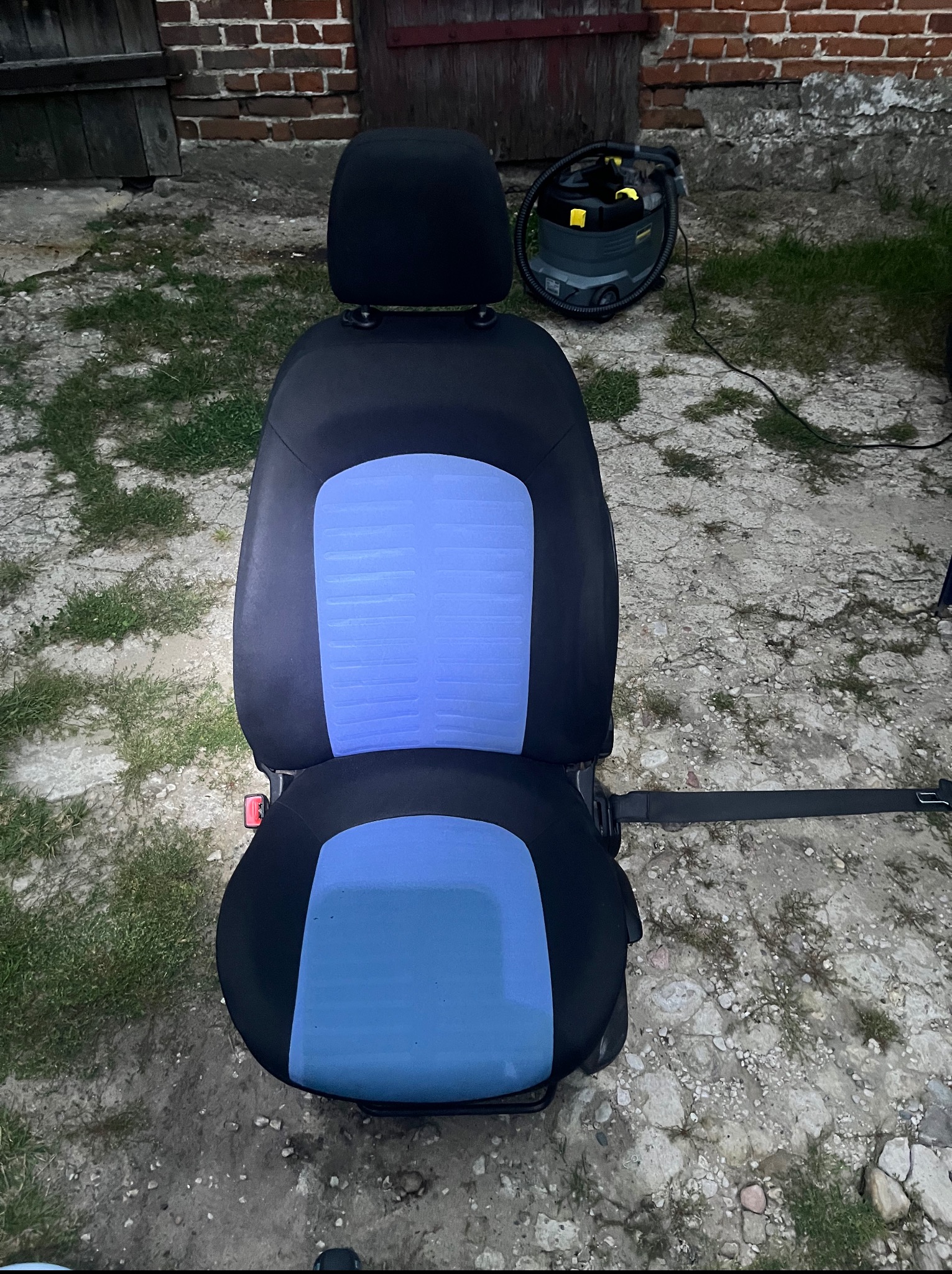 Wyjęty fotel samochodowy z niebieską tapicerką, obok odkurzacz piorący Karcher. Na zewnątrz, podłoże z kamieni i trawy. Widoczne ślady użytkowania.