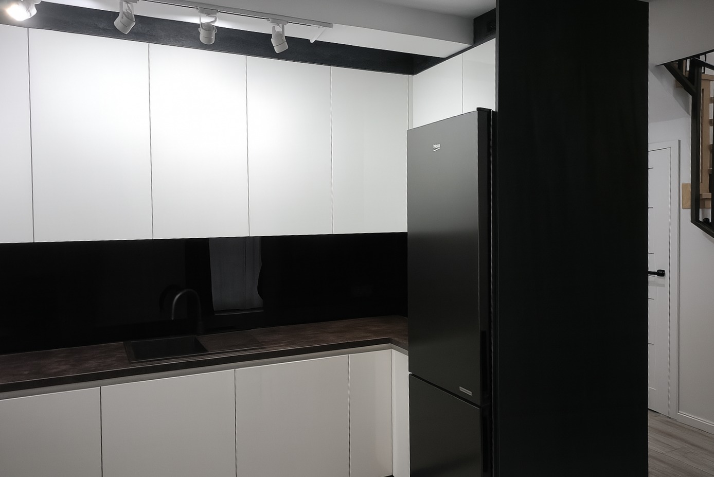 Minimalistyczna kuchnia w bieli i czerni: białe szafki, czarny panel, ciemny blat i lodówka Beko. Nowoczesne oświetlenie punktowe.