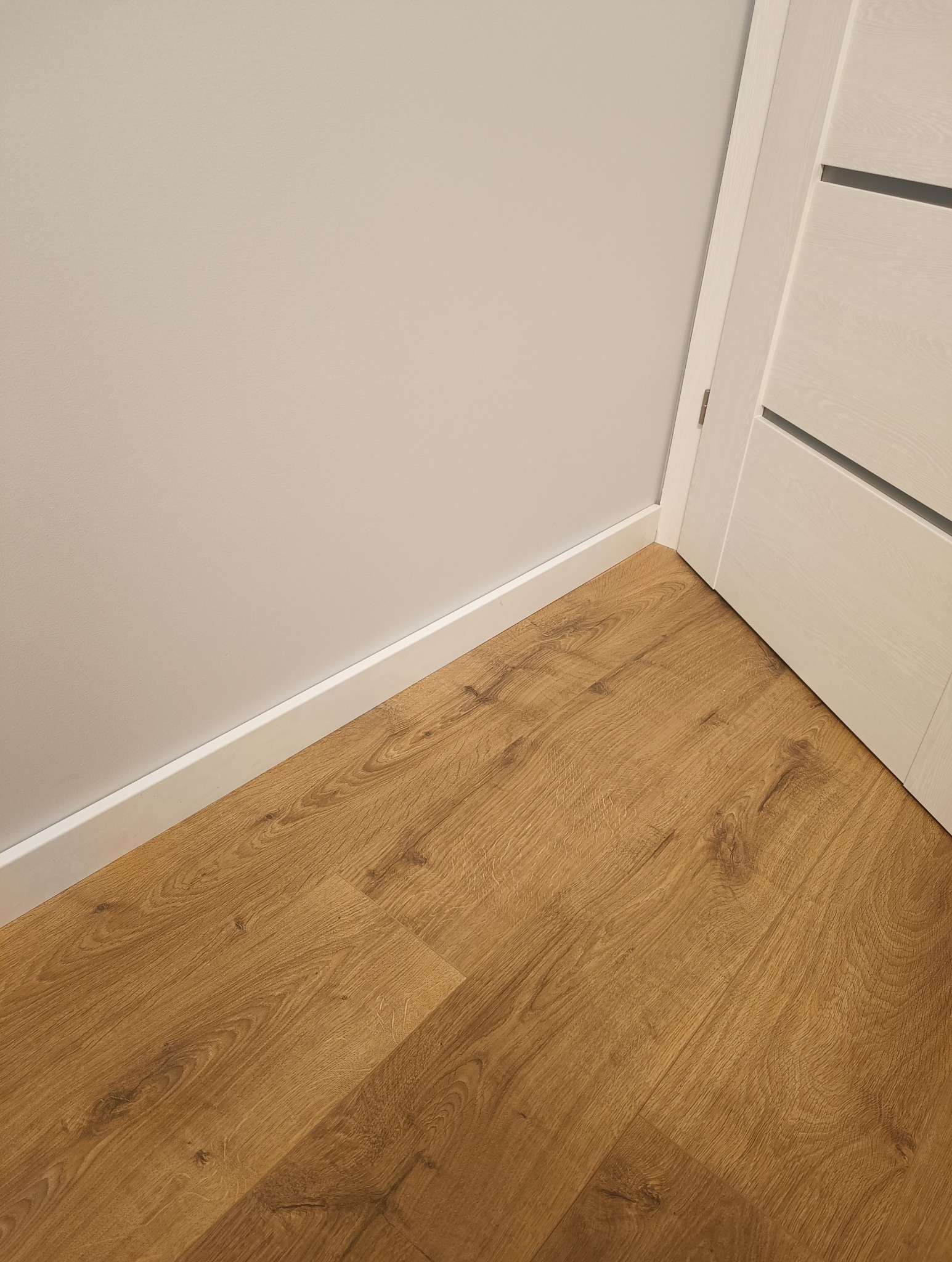 Nowa podłoga z paneli w odcieniach dębu, ułożona przy ścianie i białej listwie przypodłogowej, widok na fragment białych drzwi z szarymi wstawkami.