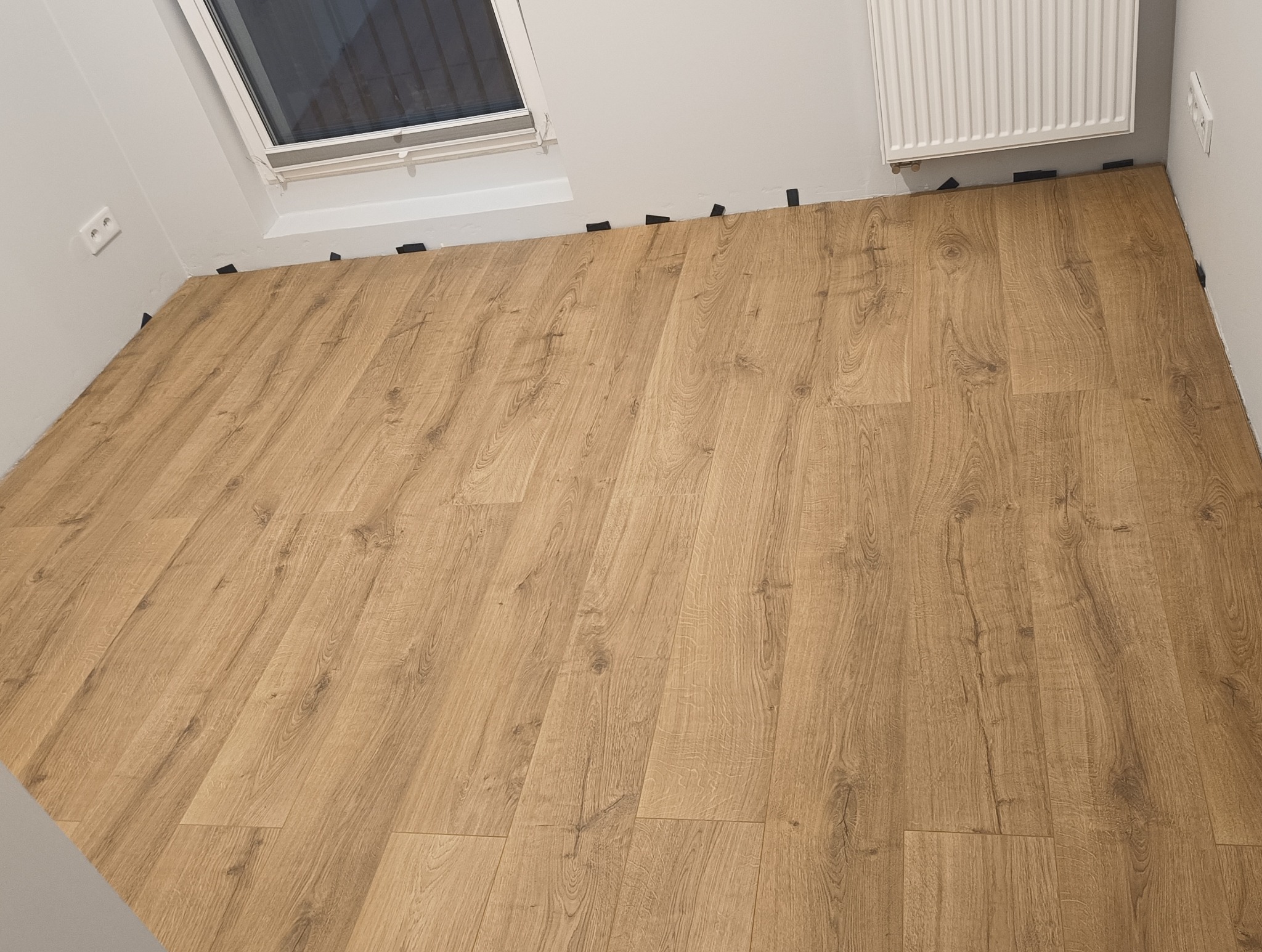 Jasne panele podłogowe ułożone w pokoju z oknem dachowym i grzejnikiem. Widoczne czarne kliny dystansowe przy ścianach. Realizacja w trakcie.