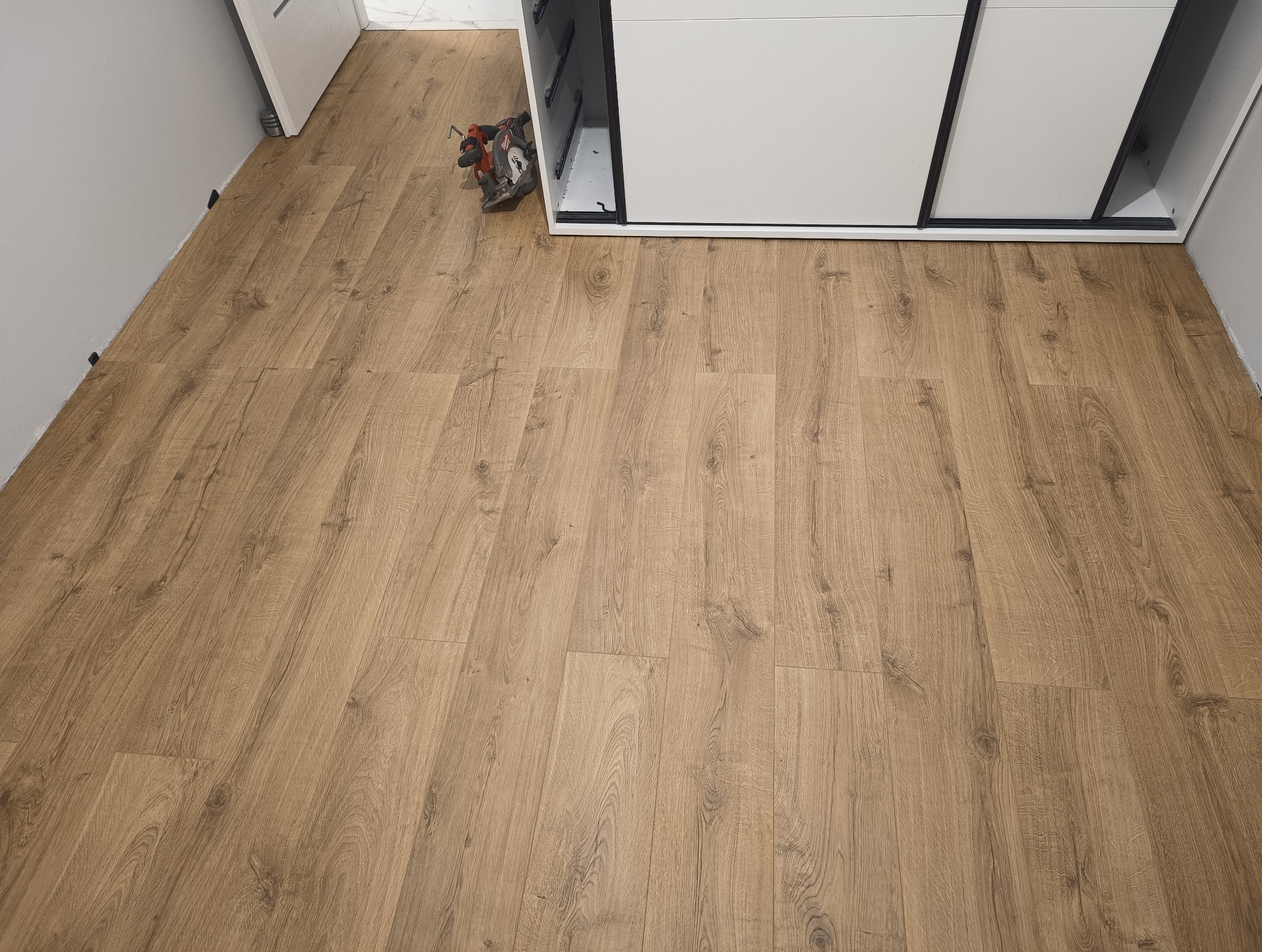 Ułożone panele podłogowe w odcieniach drewna, widoczne narzędzie do cięcia przy szafie przesuwnej. Nowa podłoga w jasnym wnętrzu.