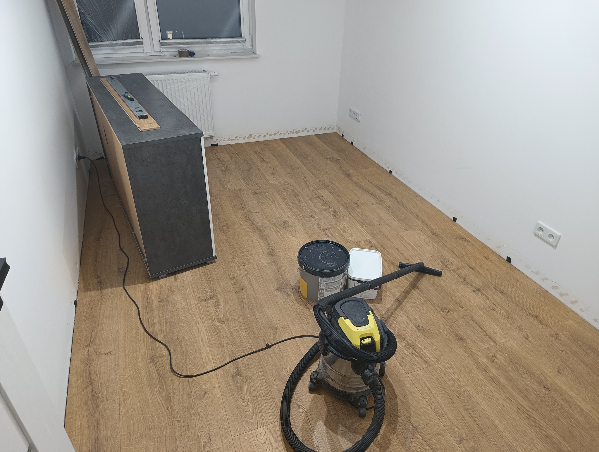 Ułożone panele podłogowe w pokoju w Pruszkowie, widoczny odkurzacz przemysłowy i wiaderka po farbie, listwy przypodłogowe jeszcze nie zamontowane.