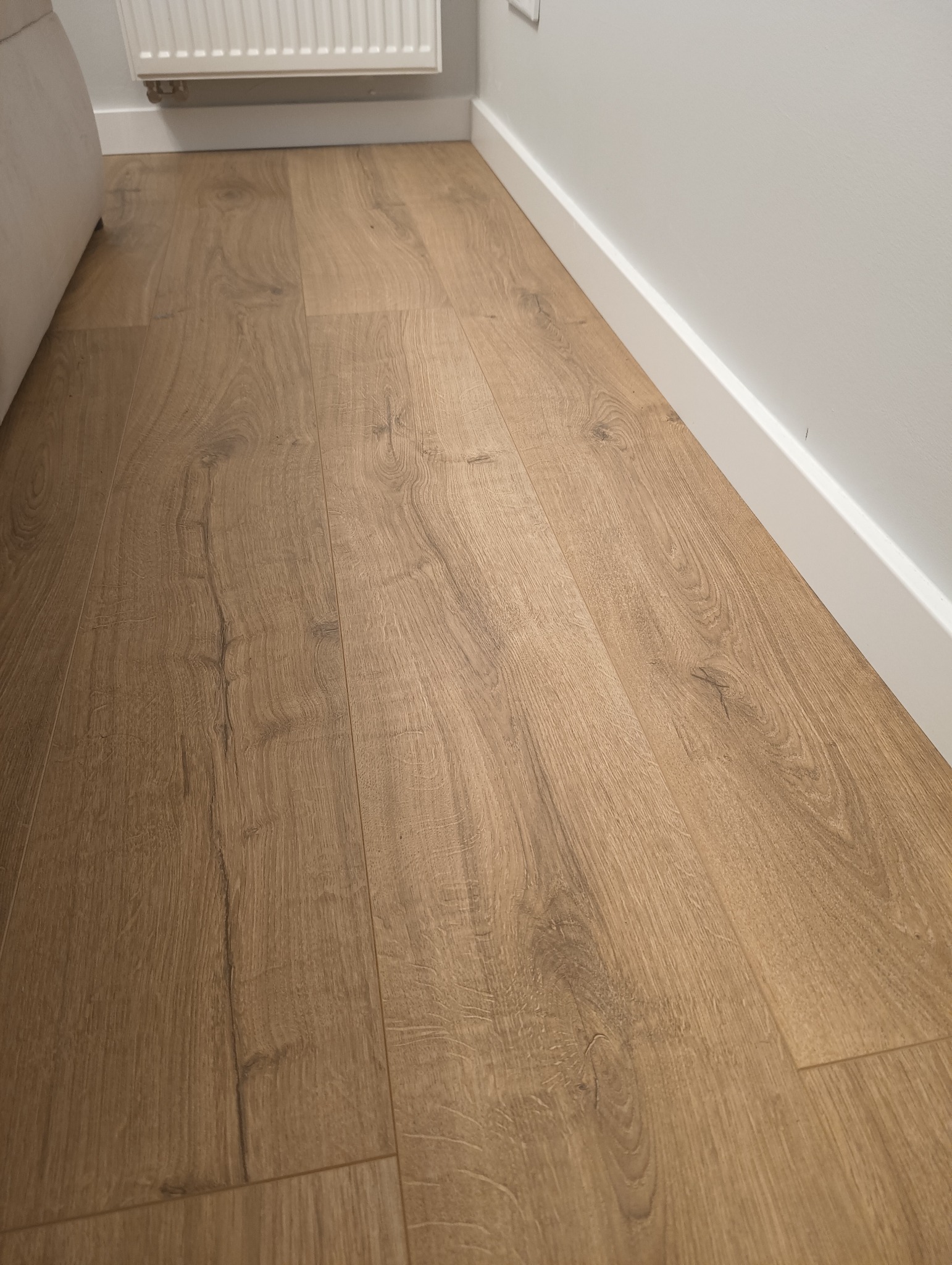Jasne panele podłogowe ułożone wzdłuż ściany z białą listwą przypodłogową. Widoczny fragment grzejnika i narożnik sofy.