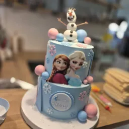 Dwupoziomowy tort urodzinowy w jasnych barwach z motywem 'Kraina Lodu', dekorowany figurką Olafa i imieniem solenizantki.