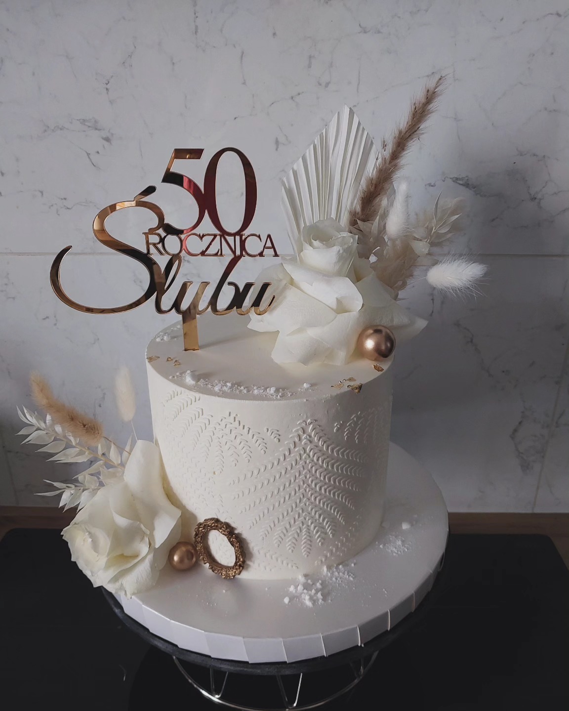 Tort na 50. rocznicę ślubu: biały, zdobiony wzorem paproci, z topperem i kwiatami. Dekoracje w odcieniach bieli i złota, elegancki design.