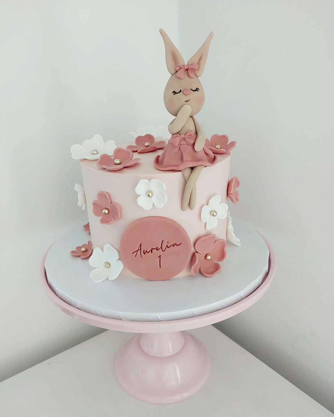 Tort na roczek w pastelowych barwach z figurką królika i kwiatami. Okrągły napis 'Aurelia 1' na różowym tle. Całość na różowej paterze.