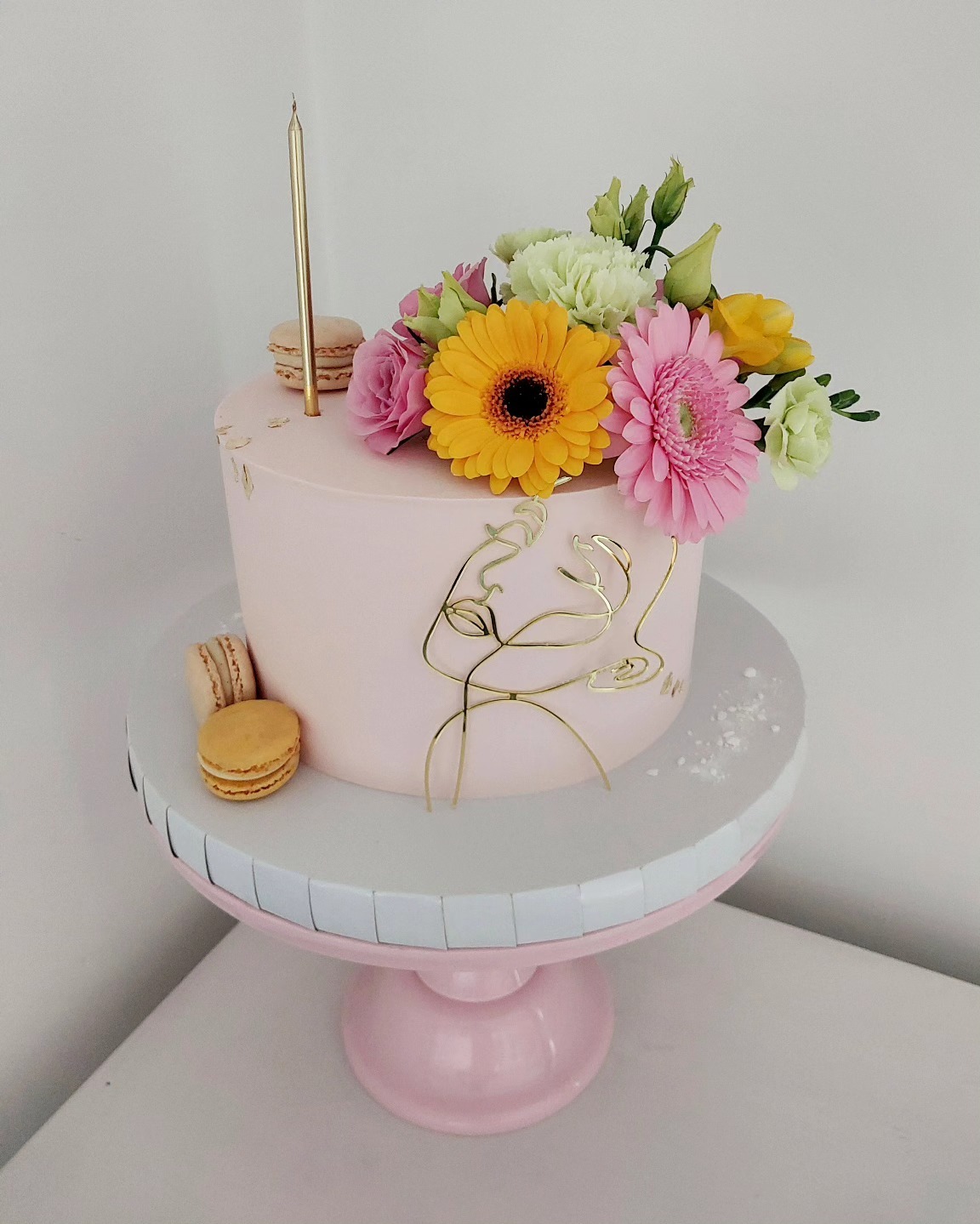 Różowy tort z kwiatami i złotą dekoracją na paterze z makaronikami, złota świeczka, minimalistyczny design.