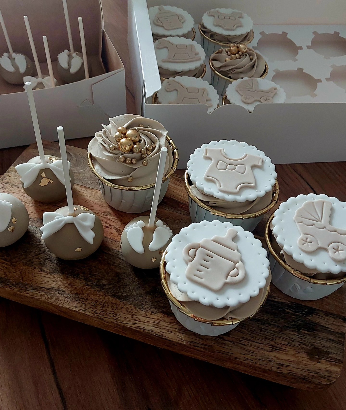 Artystyczne babeczki i cake pops z motywami dziecięcymi (butelka, wózek, ubranko) na drewnianej desce, idealne na baby shower. Kremowe dekoracje.
