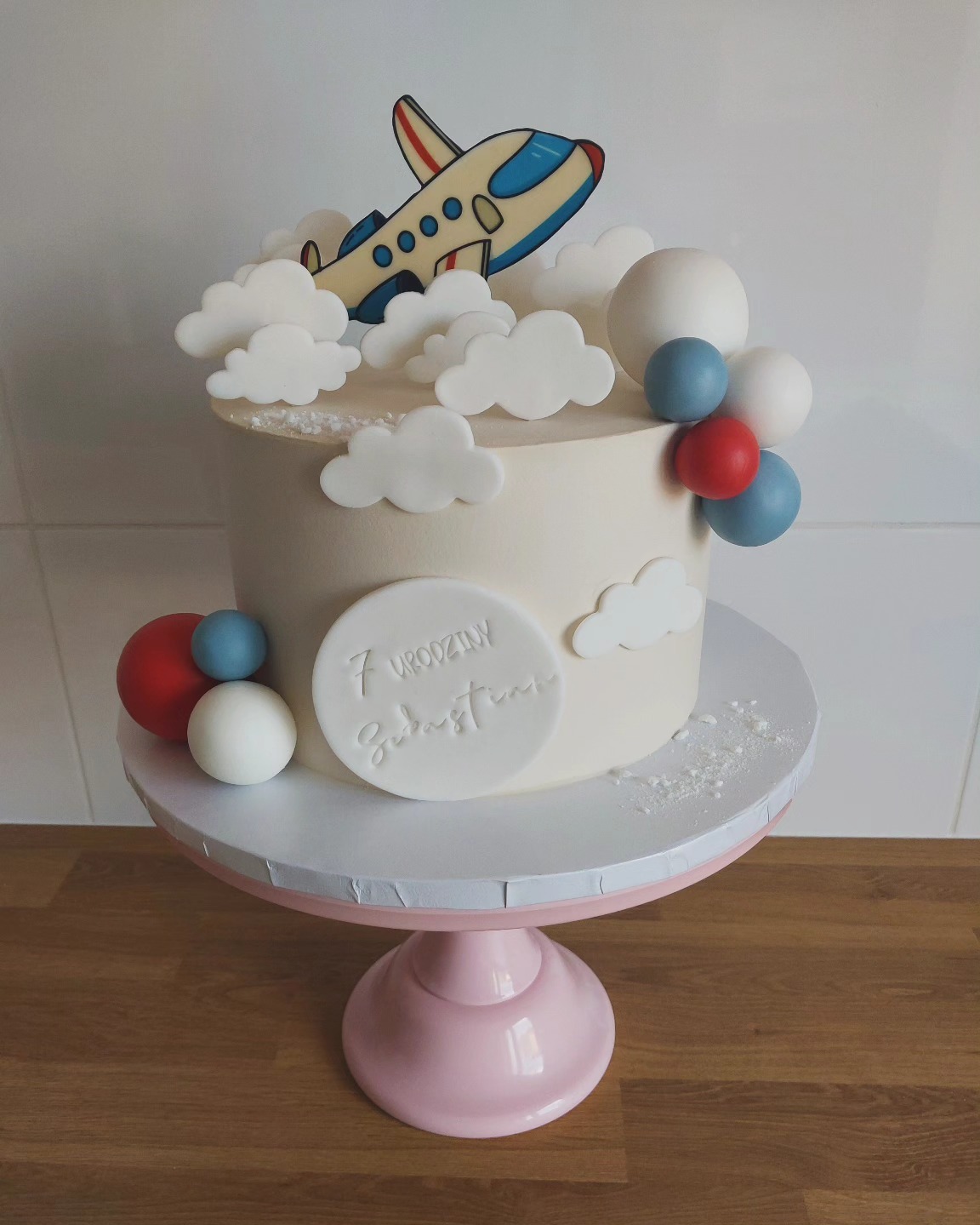 Tort urodzinowy dla 7-latka z motywem samolotu i chmur na różowym stojaku, imię 'Sebastian' wytłoczone na boku.