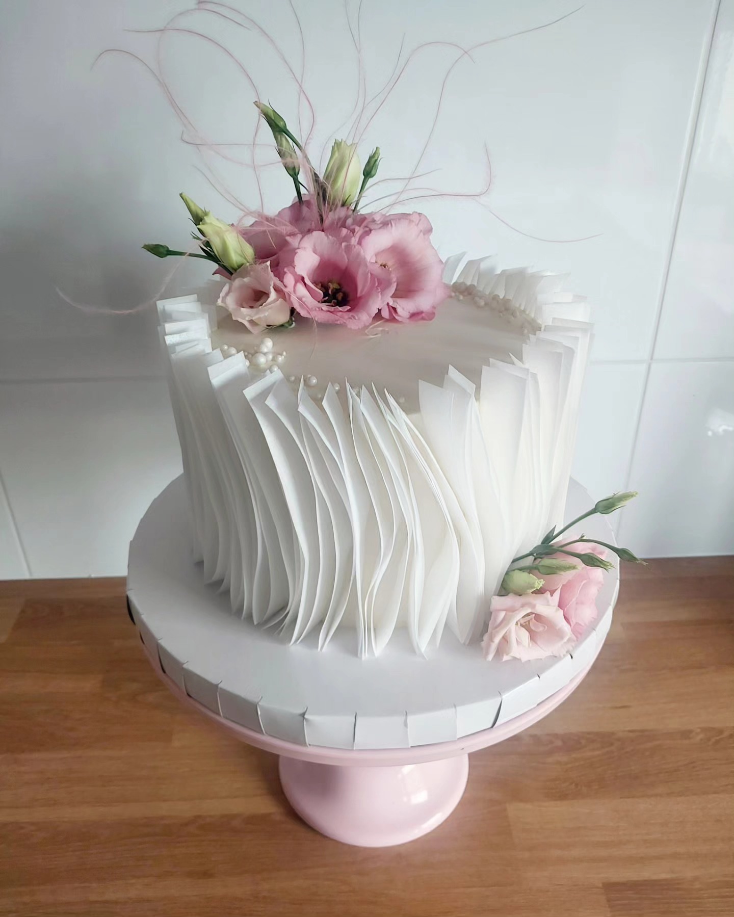 Elegancki tort z białymi, falistymi zdobieniami i różowymi kwiatami na różowej paterze. Dekoracja z piórek, minimalistyczny design, jasne tło.