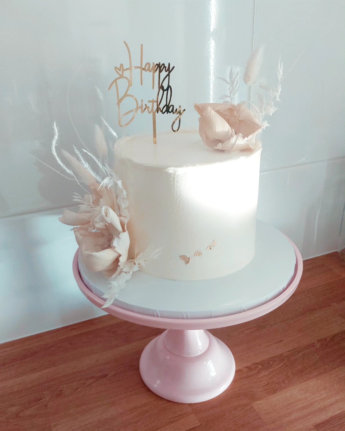 Tort urodzinowy w kremowych odcieniach, ozdobiony kwiatami i napisem 'Happy Birthday' na różowym stojaku, prezentowany na tle białej ściany.